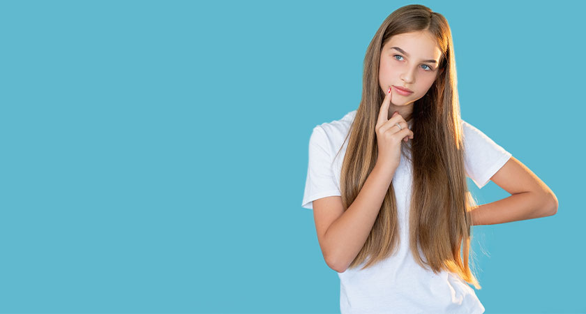 A imagem mostra uma menina branca, de cabelos castanhos, lisos e longos. Ela veste camiseta branca e está posicionada à direita da imagem. Sua mão esquerda está posicionada em sua cintura e seu dedo indicador direito, toca suavemente seu queixo. O fundo da imagem é azul.