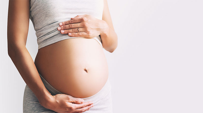 A imagem mostra uma gestante, no canto esquerdo da imagem. Ela veste blusa e calça cinzas e está com a barriga à mostra. Suas mãos estão posicionadas, uma acima e outra abaixo da barriga. O fundo da imagem é cinza.