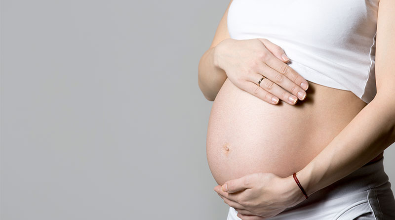 A imagem mostra uma mulher gravida, com as mãos postas sobre a barriga. Ela veste um top branco e calças brancas. A foto enquadra apenas da região do peitoral até a cintura da mulher. O fundo da imagem é cinza.