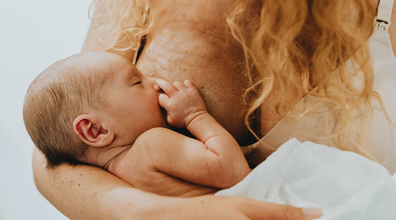 A imagem mostra o tórax de uma mulher branca com cabelos loiros e cacheados. Ela está usando um sutiã bege e, com a mama esquerda, amamenta um bebê que aparenta ser recém-nascido e está envolto em um pano branco. 