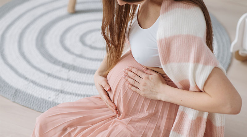 A imagem mostra uma mulher caucasiana sentada em uma cadeira. Ela veste uma regata branca, saia rosa e casaco listrado. A mulher, que está grávida, segura a barriga com ambas as mãos. Não é possível visualizar seu rosto.