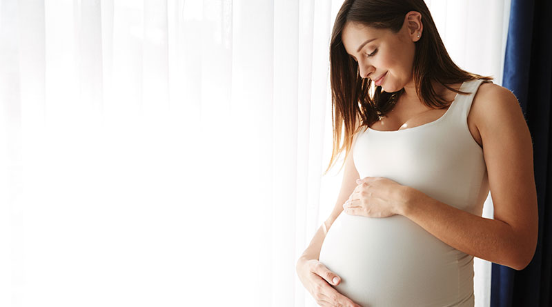 A imagem exibe uma mulher branca, gestante, de cabelos castanhos e lisos, na altura do ombro. Ela veste regata branca e com as mãos toca sua barriga, enquanto sorri. Atrás dela é possível visualizar uma cortina branca.