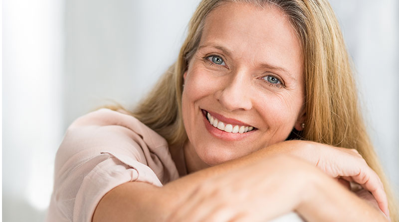 A imagem mostra uma mulher caucasiana, de meia-idade, com cabelos loiros, olhos azuis e sorridente. Ela está com os braços apoiados nos joelhos e olha para a frente.