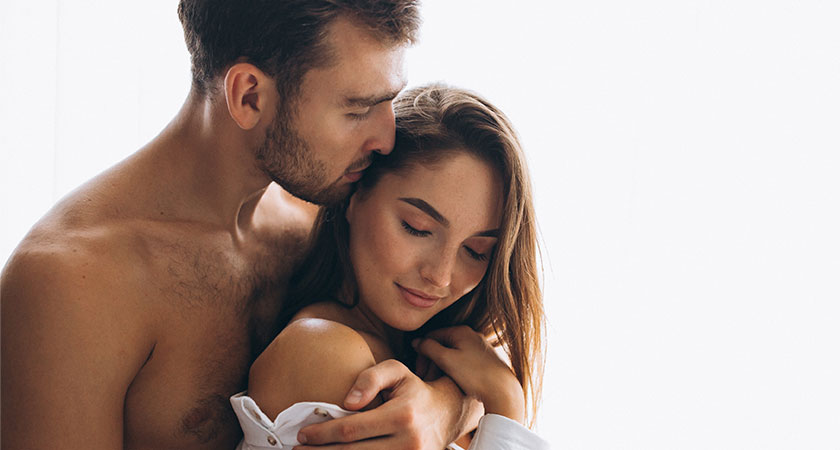 A imagem exibe um jovem casal, em frente a um fundo branco. Ele é branco, tem cabelos e barba castanhos e está sem camisa, posicionado atrás da mulher. Seu rosto está apoiado sobre a cabeça dela, e seu braço esquerdo está abraçando-a. A mulher é branca, tem cabelos lisos, curtos e castanhos, e veste uma camisa branca, com um ombro à mostra. Ela está com a cabeça levemente inclinada para frente, e sua mão direita toca o braço do homem.