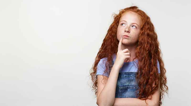 A imagem mostra uma menina jovem, de cabelos longos e ruivos. Ela está usando uma camiseta lilás e uma jardineira jeans. Seus braços estão cruzados e uma de suas mãos está próxima ao rosto, que expressa dúvida.