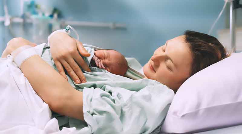 A imagem mostra um ambiente hospitalar, com paredes azuladas e equipamentos de saúde. Em uma cama, há um lençol lilás, e nela há uma mãe com seu bebê recém-nascido.