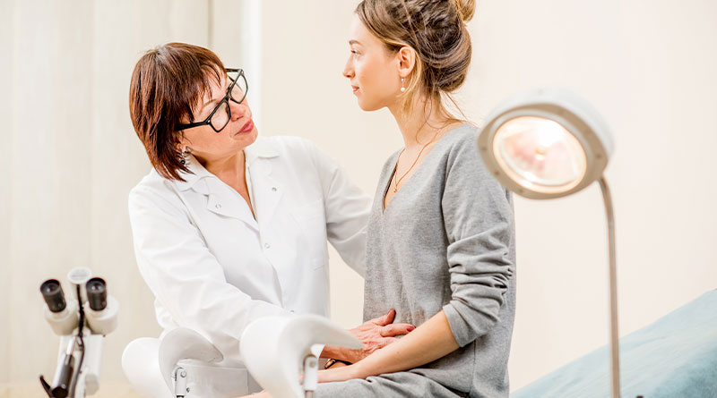 Em um consultório predominantemente branco, uma médica atende uma paciente. A médica, de jaleco branco, cabelos pretos e usando óculos conversa com uma jovem paciente, colocando a mão calmamente em seu abdômen. A paciente está com semblante atento, prestando atenção no que a médica tem a dizer.