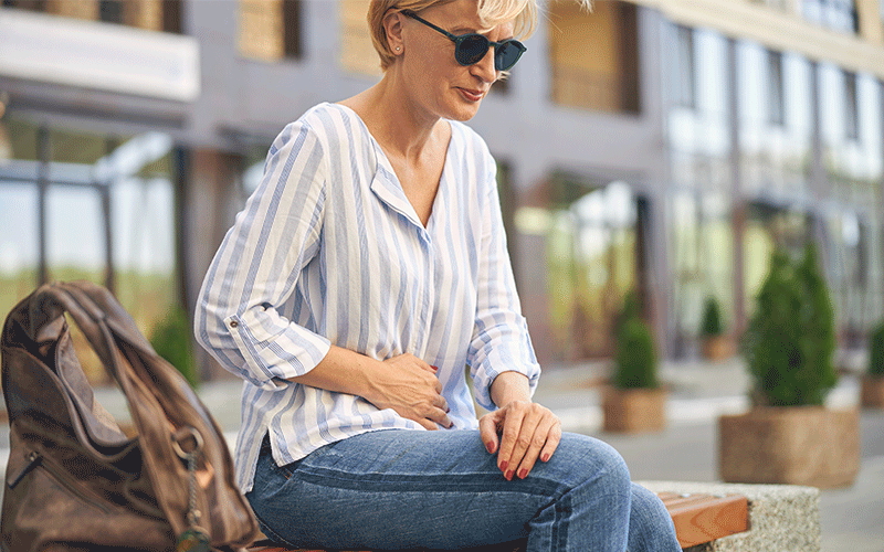 Na imagem há uma mulher loira com camisa azul e branca listrada, calça jeans e óculos escuro. Ela está sentada em um banco, em uma pequena praça com plantas aos arredores e edifícios. Ao seu lado, no banco, há uma bolsa de couro marrom. Ela está com a mão no abdômen com um semblante desconfortável.